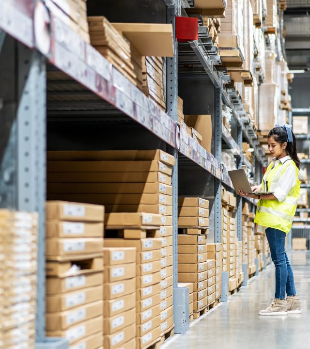 Asian engineer woman check stock details on tablet computer for checking boxes with logistics on shelves with goods background in warehouse